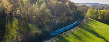 Pohled z výšky na modrý vlak jedoucí podél zelené louky na jedná straně a zeleného kopce na druhé straně.