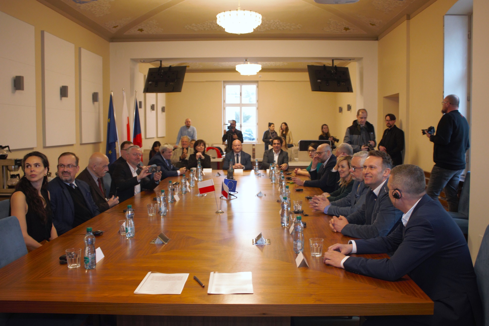 Všichni představitelé české a polské strany sedí u velkého jednacího stolu, na kterém jsou vlaječky České republiky a Polska.
